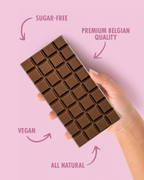Chocolate bar held by a hand with text highlighting sugar-free, premium Belgian quality, vegan, and all natural attributes on a pink background.